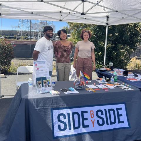 Three people standing behind a table. The tablecloth says "Side by Side" in bold letters. 